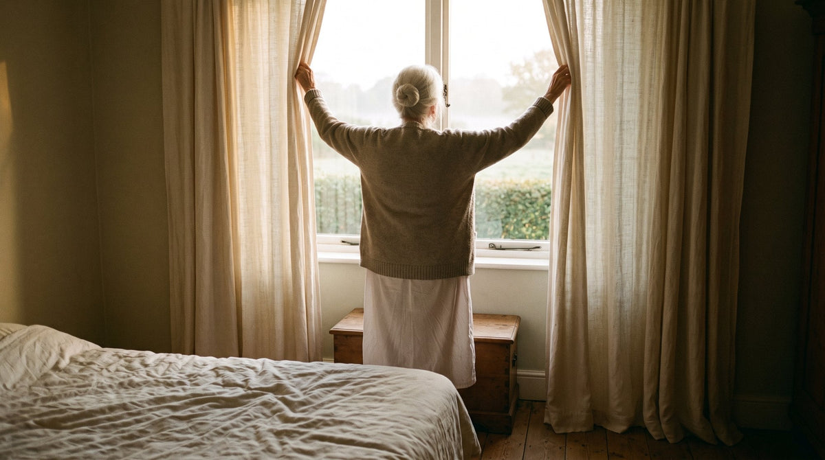 Une grand-mère ouvre les rideaux de sa chambre en levant les bras, au lever du jour