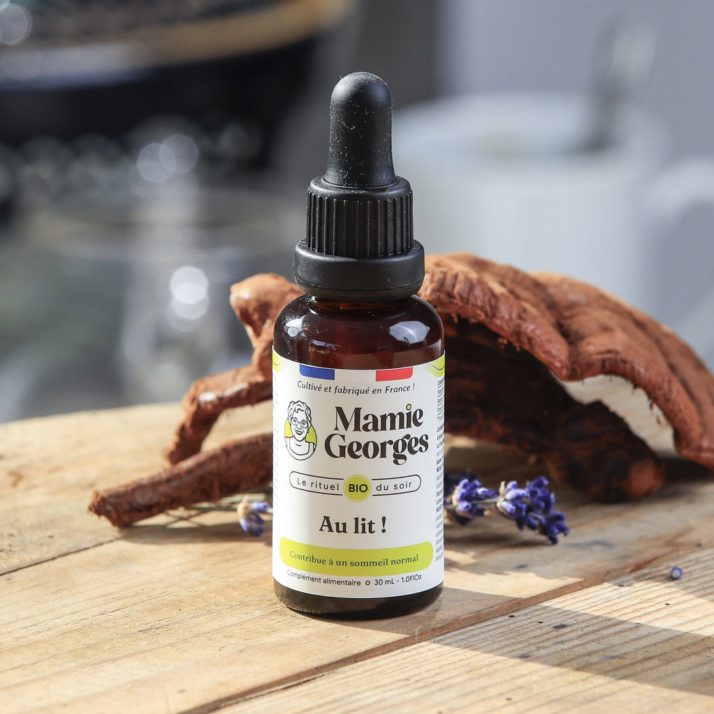 Flacon Au lit ! de Mamie Georges, fermé, sur une table en bois devant un reishi, une tasse blanche en arrière-plan