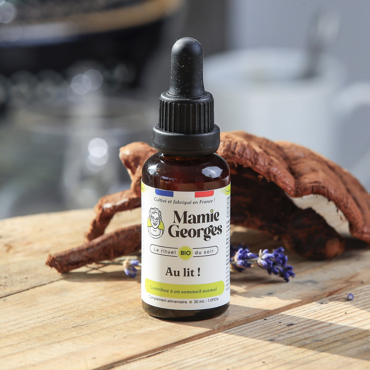 Flacon Au lit ! de Mamie Georges, fermé, sur une table en bois devant un reishi, une tasse blanche en arrière-plan
